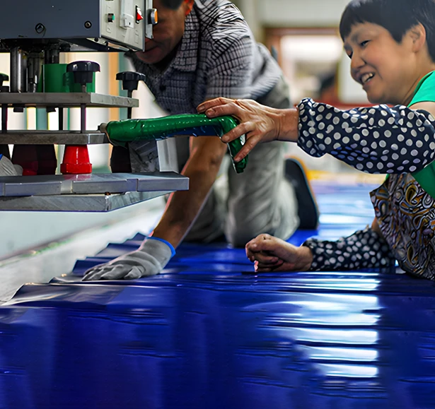 Two people are working together with machinery, handling a large sheet of blue material in a workshop setting.