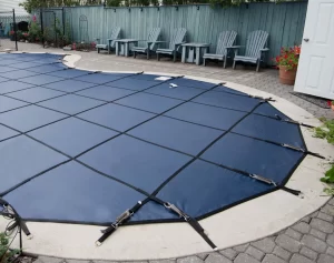 An in-ground pool covered with a blue safety cover, surrounded by a paved patio area and lined with several outdoor chairs against a wooden fence.