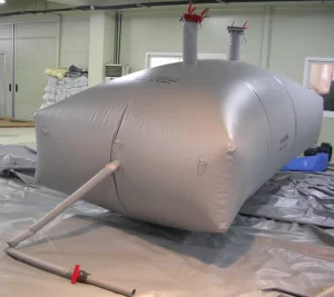 An inflated, rectangular grey plastic tank with two vertical pipes on top and a hose attached on the side, placed on a grey sheet inside an indoor facility.