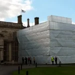 A large section of a historic building is covered in Clear String Reinforced Poly Tarps – 7 oz FR Scaffold Sheeting for renovation work. Several people are standing near the covered area, and a flag is flying atop the building.