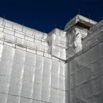 A building under construction is fully covered with Clear String Reinforced Poly Tarps – 7 oz FR Scaffold Sheeting beneath a clear blue sky, with scaffolding visible along the top edges.