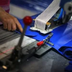 A person's hand operates a machine handling blue fabric, with part of the Custom Truck Tarp, Trailer Tarp machinery and fabric rolls visible in the foreground.