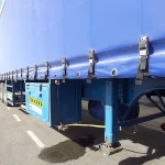 A blue semi-trailer truck with a Custom Truck Tarp, Trailer Tarp cover is parked on the side of a road. Multiple black buckles secure the tarp in place along the side of the trailer.