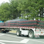 A blue semi-truck with a Lumber Tarp for Flatbed Truck and Trailer covering its long flatbed trailer is driving on a road bordered by tall pine trees.