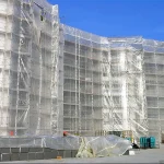 A large building under construction is covered in FR SCAFFOLD SHEETING-10oz, with construction materials and equipment scattered around the site.