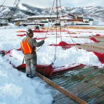 A worker in an orange safety vest operates lifting equipment to move Snow Tarps filled with snow at a construction site surrounded by snow-covered mountains.