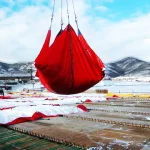 A crane suspends a large red container equipped with Snow Tarps over a snowy, industrial area with mountains in the background.