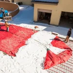 Workers use Snow Tarps to clear snow from a construction site in front of a house with open garage doors. Snow is piled on the tarps, and one worker holds a rope to drag the tarp.